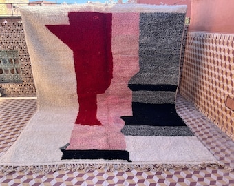 May include: A white rug with a geometric design in shades of pink, red, black, and gray. The rug has a fringe border.