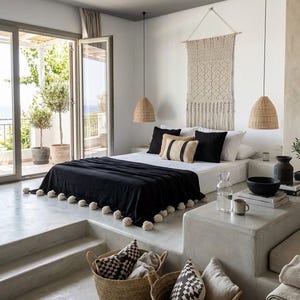 Op de afbeelding: Slaapkamer met een minimalistisch design. Het bed heeft een zwarte deken met witte pompons en decoratieve kussens. Twee geweven hanglampen hangen aan het plafond. Twee geweven manden met kussens op de voorgrond.