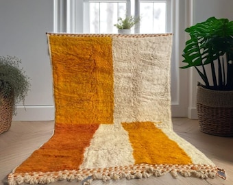 May include: A rectangular, hand-woven rug with a geometric design. The rug features a color block pattern with sections of orange, cream, and rust. The rug has a fringed edge and is displayed on a wooden floor.
