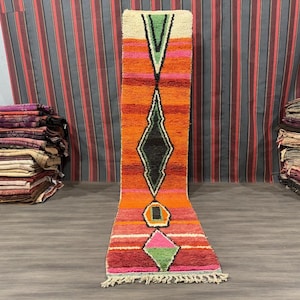 Puede incluir: Una alfombra larga y estrecha con un patrón geométrico en tonos naranja, rosa, verde y negro. La alfombra tiene flecos en ambos extremos.
