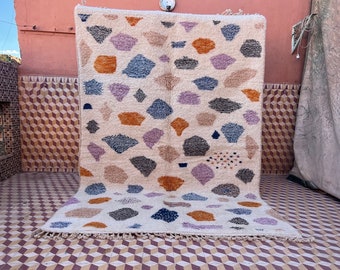 May include: A white rug with a geometric pattern of colorful shapes, including orange, blue, pink, and gray. The rug has a fringe border and is laid out on a tiled floor.