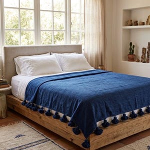 May include: A bed with white linens and a navy blue throw blanket. The blanket has a textured appearance and is adorned with navy blue tassels along the edge. The bed frame is made of light-colored wood, and the room has a neutral color palette.