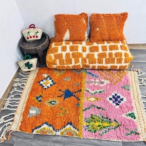 May include: An orange and pink rug with a geometric design, paired with orange pillows and a long cushion. Two small woven baskets with fruit and cactus designs sit on a wooden stool. The rug has a fringed edge.