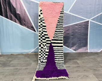 May include: A long, rectangular rug with a geometric design. The rug features a pink triangle at the top, a black and white checkered pattern, and a purple triangle at the bottom. The rug has a white background with black stripes.