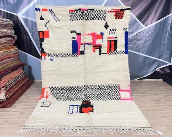 May include: A white Moroccan rug with a geometric pattern in black, red, pink, and blue. The rug has a fringe border.