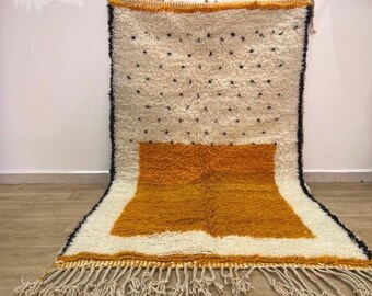 May include: A white and yellow Moroccan rug with a geometric pattern and fringe. The rug has a white background with a yellow square in the center and black dots scattered throughout.