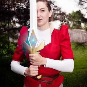 Puede incluir: Una mujer con un traje rojo y blanco sostiene una espada grande, dorada y blanca con una gema azul en el centro. La espada tiene una hoja blanca y una empuñadura dorada.