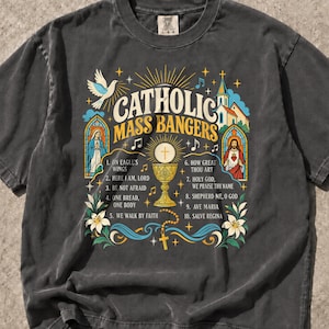 May include: A charcoal gray short-sleeve t-shirt featuring a graphic print with the text Catholic Mass Bangers, a golden chalice, a dove, religious figures in stained glass windows, and a list of ten song titles including On Eagle's Wings.