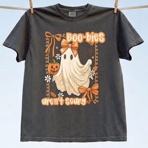 May include: A charcoal gray t-shirt featuring a graphic of a ghost wearing an orange bow, holding a jack-o'-lantern, and surrounded by orange bows and flowers. The text reads "Boo-bies aren't scary" in orange lettering with a decorative border.