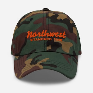 May include: Camouflage baseball cap with the text "Northwest Standard Time" in orange. The cap has a curved peak and a black button. The pattern includes shades of green, brown, and tan.