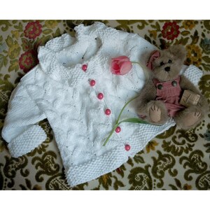 May include: White knitted cardigan with a ruffled collar and pink buttons. The cardigan is laid out on a floral patterned fabric with a teddy bear and a pink tulip.