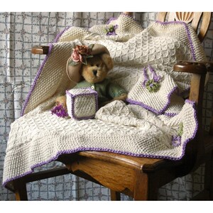Könnte beinhalten: Eine weiße gestrickte Babydecke mit einem violetten Rand und einem Teddybär, der einen Hut und ein grünes Hemd trägt. Die Decke liegt auf einem Holzstuhl mit einem floral gemusterten Stoffhintergrund.