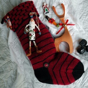 Könnte beinhalten: Ein rot-schwarzer gestrickter Strumpf mit schwarzem Bündchen und einem weißen und roten Ornament, das daran hängt. Der Strumpf liegt auf einem weißen Stoffhintergrund mit anderen Weihnachtsdekorationen.