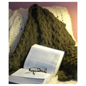 May include: A green knitted throw blanket draped over a white crocheted throw pillow and a book with reading glasses on top.