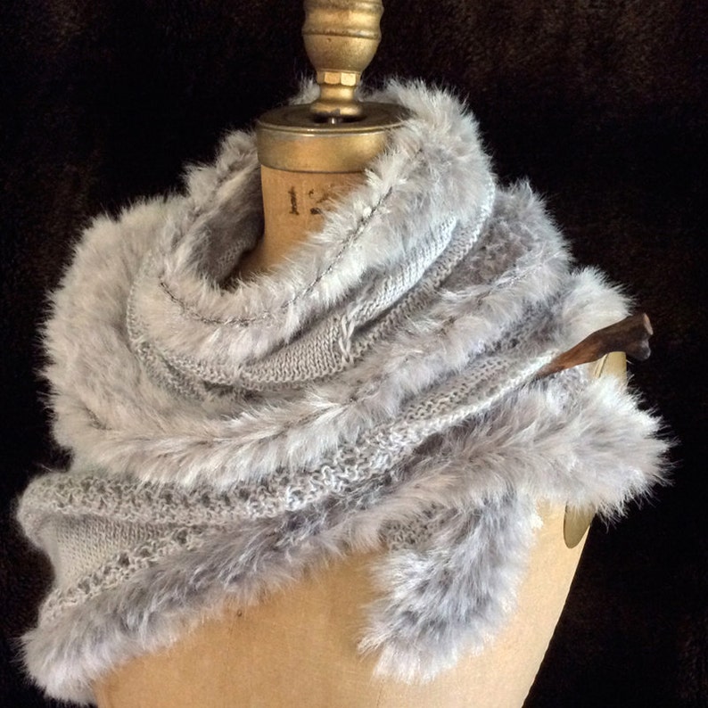 May include: A gray knitted scarf with a faux fur trim. The scarf is draped over a mannequin's shoulders.
