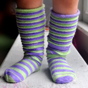 Könnte beinhalten: Ein Paar gestreifte Kniestrümpfe in Grün- und Lilatönen. Die Socken sind aus weichem, gestricktem Stoff und haben eine bequeme Passform.