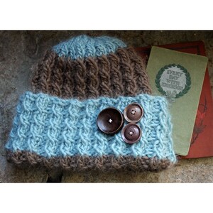 May include: A brown and blue knitted hat with three brown buttons. The hat has a cable knit pattern and is folded down at the brim.