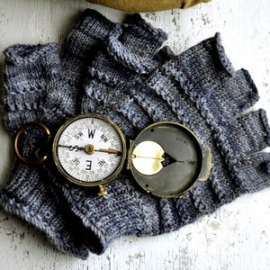 Könnte beinhalten: Ein grauer, fingerloser Strickhandschuh mit einem Kompass darauf. Der Kompass hat ein Messinggehäuse und ein weißes Zifferblatt mit schwarzen Markierungen.