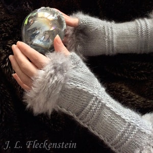 May include: A pair of gray knitted fingerless gloves with a fluffy white trim. The gloves are held in a person's hands, and a clear glass ball is in the background.