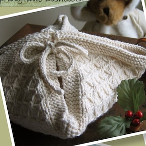 Peut inclure: Un panier en tricot blanc avec un nœud, réalisé avec un motif de point de câble. Le panier est posé sur une surface en bois avec une feuille verte et des baies rouges.