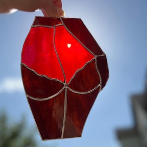 May include: A stained glass art piece in a geometric shape, featuring shades of red and burgundy. The glass is segmented with silver-colored metal. The sun shines through the glass, creating a bright spot in the center. The artwork is held up against a blue sky.