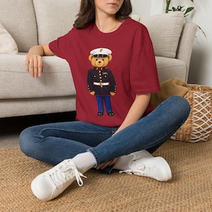 May include: A maroon short-sleeved t-shirt featuring a graphic of a teddy bear dressed in a formal dark blue military uniform with a white peaked cap, gold buttons, and blue trousers, paired with dark blue denim jeans and white sneakers.