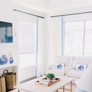 May include: A bright, airy living room with white walls and furniture. White curtains with blue trim frame a large window. Two striped armchairs with monogrammed pillows face a light wood coffee table. Woven baskets and decorative vases add texture.
