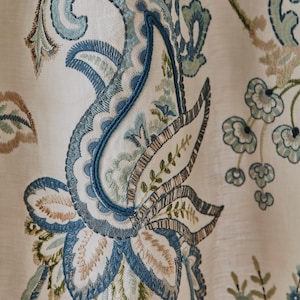 May include: Close-up of a cream-colored fabric with intricate embroidered paisley patterns. The embroidery features shades of blue, green, and tan, creating a detailed floral design. The fabric has a textured appearance, suggesting a linen or similar material.