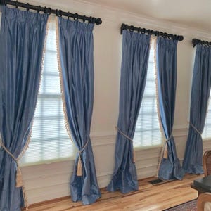 Puede incluir: Tres juegos de cortinas azules con ribete de flecos blancos y borlas que cuelgan de una ventana. Las cortinas están atadas con una cuerda.