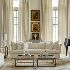 May include: A cream-colored sofa with several white pillows sits in front of a window with cream-colored curtains. Two square, dark metal coffee tables are in front of the sofa. Two decorative vases are on one table.