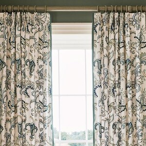 May include: Two patterned drapes with a floral design in shades of blue, green, and beige hang from a wooden rod. The drapes frame a window, creating a decorative and functional element for the room.