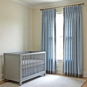 May include: A light gray wooden crib with vertical slats and a white mattress, positioned next to a window featuring floor-length light blue curtains with a subtle grid pattern and a dark gray border, hung on a metallic curtain rod with rings.