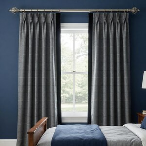 May include: A pair of floor-length pinch pleat curtains in a dark gray plaid pattern with solid black vertical side borders. The curtains are hung on a metallic silver curtain rod with decorative finials, positioned over a window with white trim.
