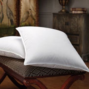 May include: Two white, fluffy pillows rest on a decorative bench. The pillows have a square shape and a textured surface. The bench has a dark, patterned cushion and a dark wooden frame with curved legs. The background includes a cabinet and a framed artwork.
