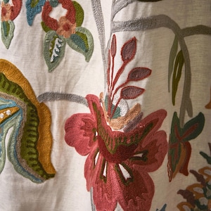 May include: Close-up of embroidered fabric with a floral design. The embroidery features a variety of colors, including red, green, blue, and yellow, on a cream-colored background. The design includes flowers and leaves.