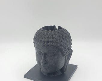 May include: A dark grey Buddha head planter, 3D-printed, with a square base. The head features detailed facial features and a textured, bubble-like hairstyle. The planter has an opening at the top for plants or decorative items.