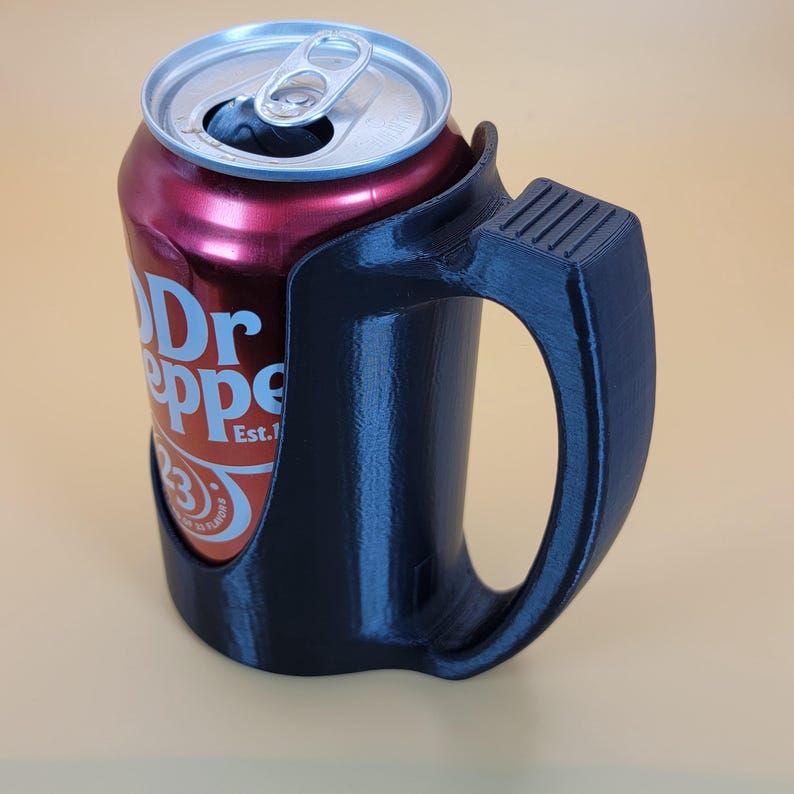 May include: A black 3D printed can holder with a handle. The holder is designed to fit a standard 12-ounce can. A red can of Dr Pepper is in the holder.