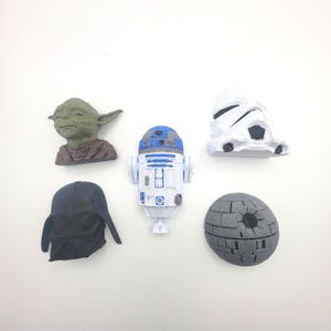 May include: A selection of Star Wars character figurines. The collection includes Yoda, R2-D2, a Stormtrooper helmet, Darth Vader's helmet, and the Death Star. The figurines are in various colours, including green, white, blue, and grey.