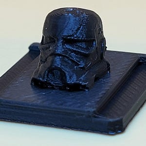 May include: A black plastic 3D printed model of a Star Wars Stormtrooper helmet on a square base.
