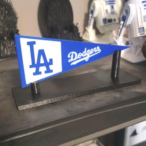 Könnte beinhalten: Ein blau-weißes Wimpel mit dem Text "LA Dodgers" auf einem schwarzen Sockel. Der Wimpel zeigt das Logo des Teams und wird von zwei schwarzen Pfosten getragen. Der Wimpel ist etwa 15 cm lang.