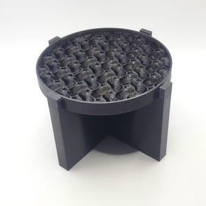 Puede incluir: Un objeto negro y redondo con un patrón de panal, posiblemente un difusor o soporte, con una parte superior circular y tres patas de soporte. El objeto está hecho de un material oscuro y se encuentra sobre una superficie blanca. El diseño presenta un patrón repetitivo de celdas interconectadas.