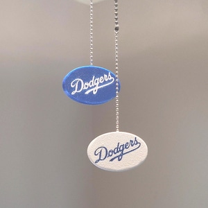 May include: Two oval-shaped fan pull chains, one blue and one white, each with the word "Dodgers" in script. The chains hang from the top of the image, suggesting a ceiling fan accessory.