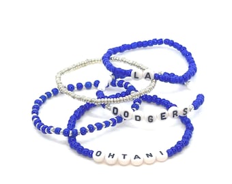 May include: Several beaded bracelets in blue and silver. Some bracelets spell out "LA", "DODGERS", and "OHTANI" with white letter beads. The bracelets are on a white background.