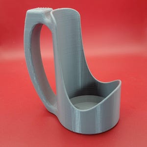 May include: A gray 3D printed cup holder with a handle. The holder is designed to hold a cup or mug.