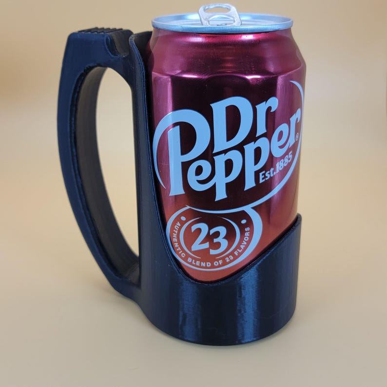May include: A black 3D printed can holder with a handle. A red can of Dr Pepper is inside the holder. The can has the text "Dr Pepper Est. 1885" and "Authentic Blend of 23 Flavors" printed on it.
