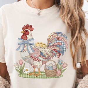 Könnte beinhalten: Cremefarbenes T-Shirt mit einem bunten Hahn-Design mit Küken, Blumen und einem Korb mit Ostereiern. Der Hahn hat einen roten Kamm und eine blaue Schleife. Das Design ist auf einem grün-weiß karierten Muster angeordnet.