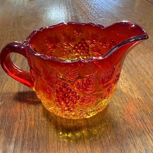 May include: An amber-colored glass creamer with a handle and spout. The creamer features a grape and leaf pattern embossed on the exterior. The glass has a textured appearance and sits on a wooden surface. A vintage kitchenware item.