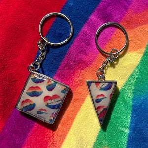 May include: Two keychains with silver-colored metal rings and chains. One keychain is square, the other is triangular. Both have a white background with a pattern of rainbow-colored lips. The keychains are displayed on a rainbow-striped background.