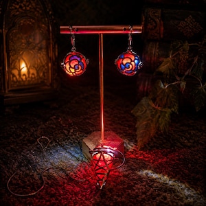 May include: A pair of spherical earrings featuring a stained-glass style rose pattern in vibrant red and blue hues, suspended from a metal display stand. A matching red, cone-shaped pendant with a geometric metal cage design rests at the base.