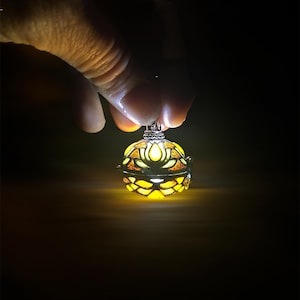 Könnte beinhalten: Leuchtender, kugelförmiger Anhänger mit floralem Design. Das warme gelbe Licht erhellt die filigranen Details vor dunklem Hintergrund. Das Design erinnert an eine Lotusblume.